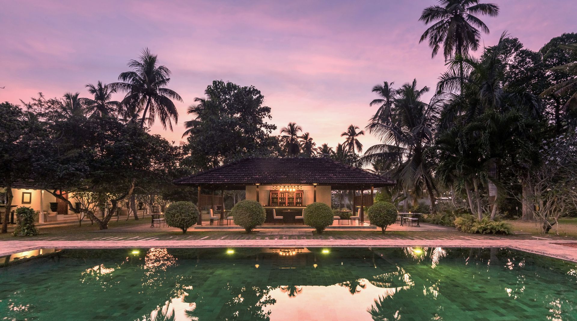 The pool at dusk under a pink sky, the lit house and palms reflected in the water
