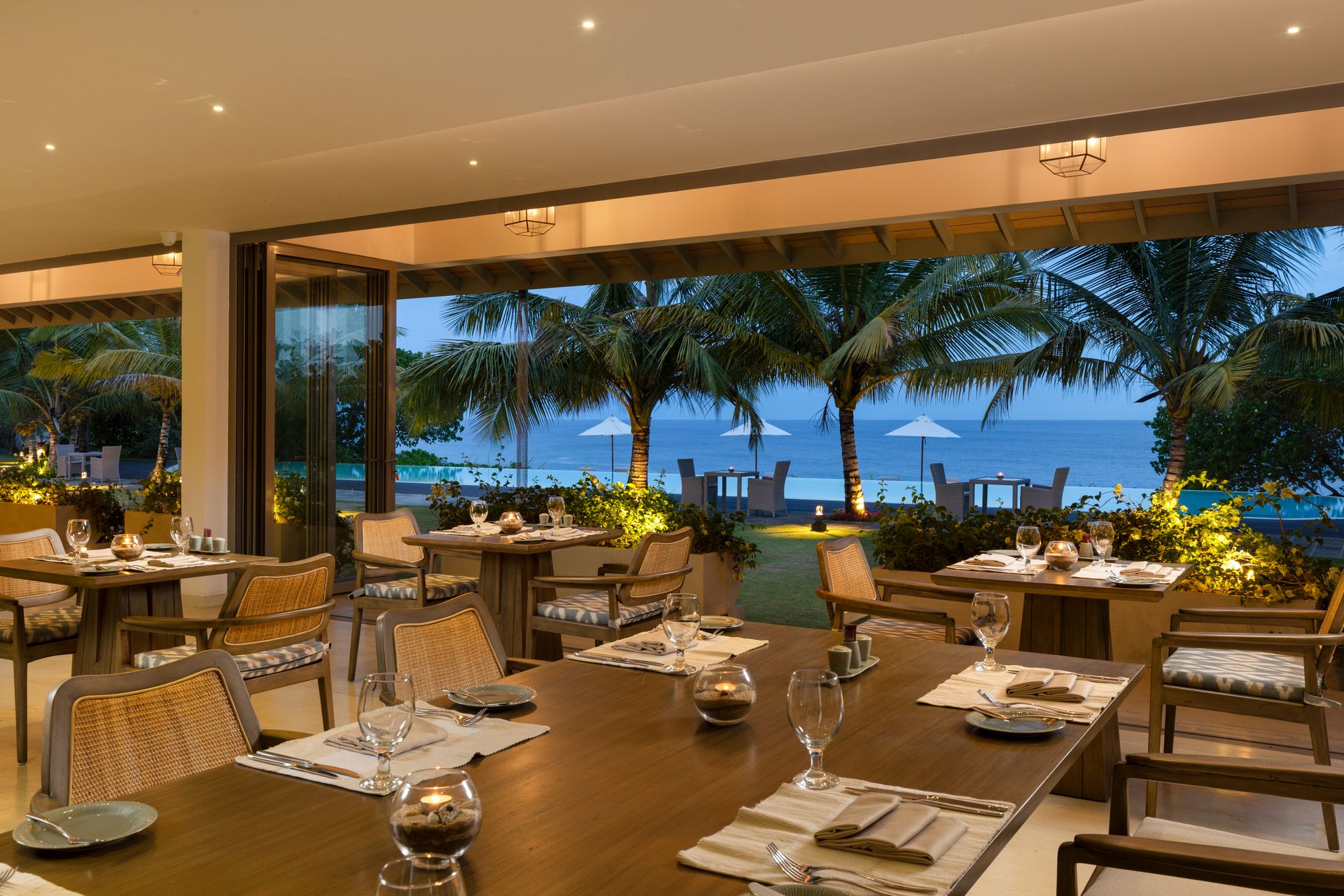 The restaurant at dusk: tables laid under a lit ceiling, open to a lawn, palms, the pool and the sea